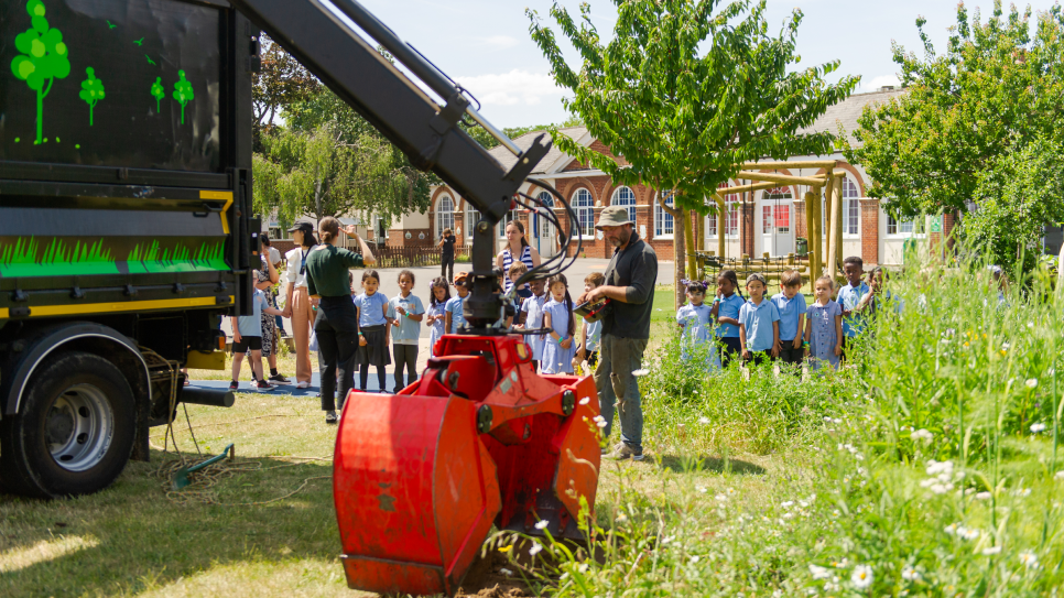 A digger lifting a tree with a group of schoolchildren in the background