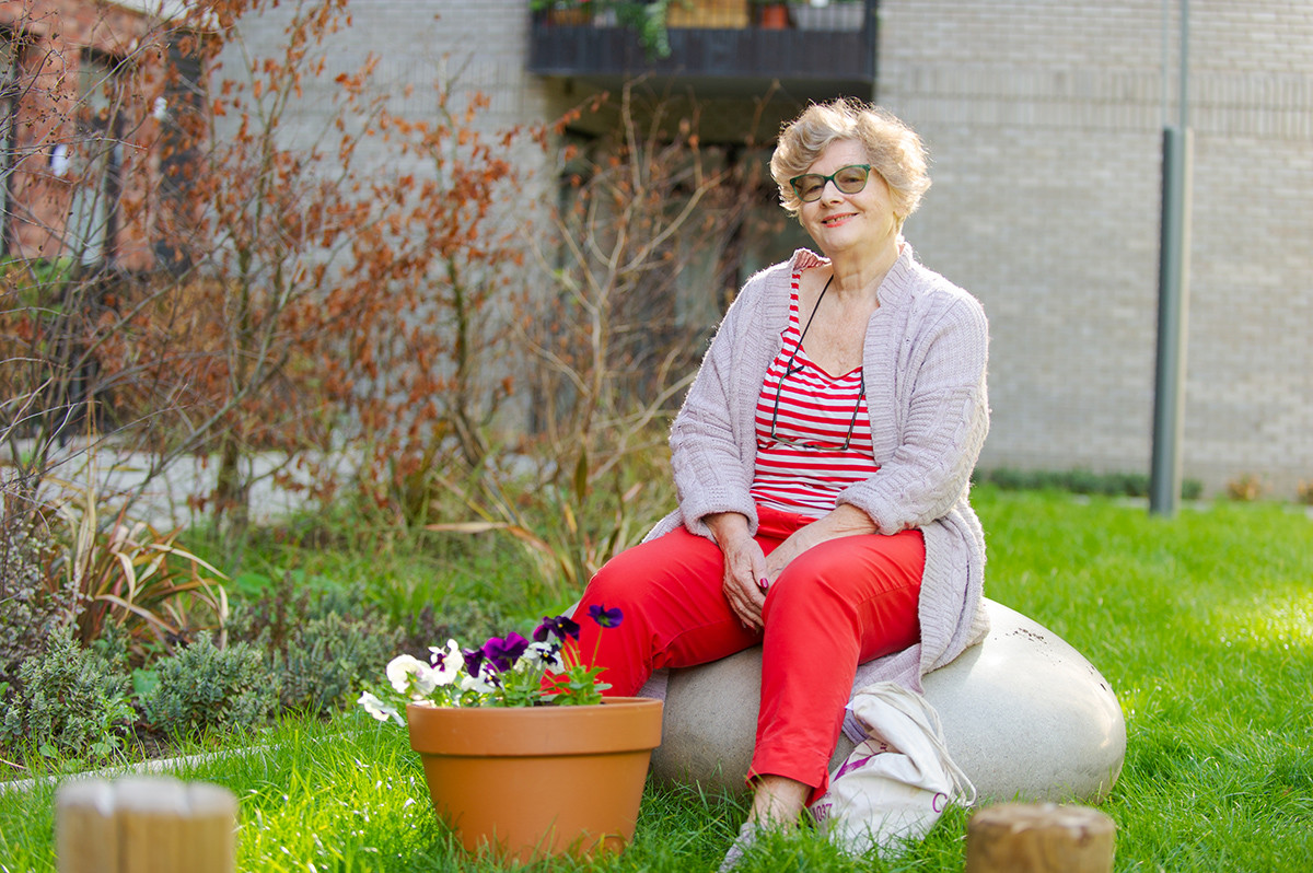 A woman sits on a boulder in a communal garden space, with some flowerpots in front of her with pansies in.